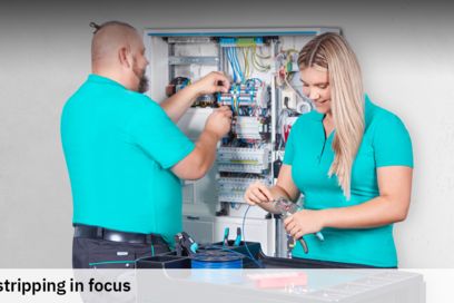 A man and a woman wearing teal-colored polo shirts are working in a control cabinet, using tools to strip wires.