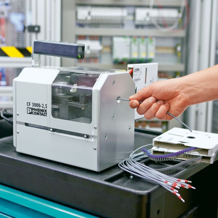 Photo of a small grey stripping and crimping machine with the Phoenix Contact logo. A hand is inserting a wire into a hole in the side of the machine, and control cabinets can be seen in the background.