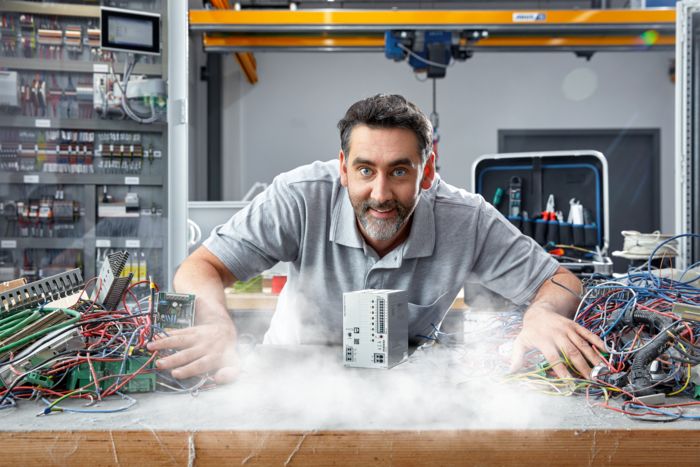 A man in a grey polo shirt pushes apart messy piles of wires to reveal a Phoenix Contact TRIO POWER supply unit. A control cabinet is behind him to the left, and to the right is a toolkit. Fog billows from the power supply unit for dramatic effect.
