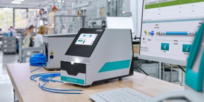 Photo of an E.FOX S 10 automatic sripping device with a touch display, sitting on a desk beside a keyboard and monitor, with a factory floor in the background.