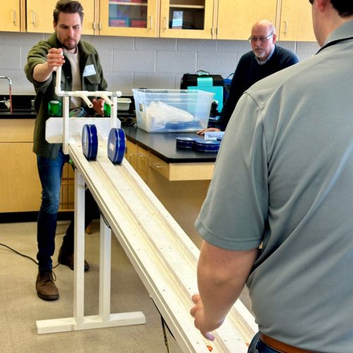 Three adult men rolling cookie tins down a makeshift ramp in a classroom