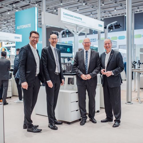 A bright photo of four men in business suits standing in front of a Phoenix Contact booth at Hannover Messe 2024. The Phoenix Contact teal and green signal chain is featured above the booth, and other people in suits can be seen walking in the background.