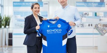 Eine Frau und ein Mann lächeln in die Kamera und halten ein Trikot von Arminia Bielefeld in den Händen